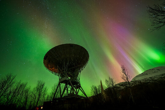 Ramfjordbotn, Troms, Norway - 07 October 2024: View of the EISCAT Tromso radar dish illuminated by vibrant green and purple aurora borealis in the night sky.
