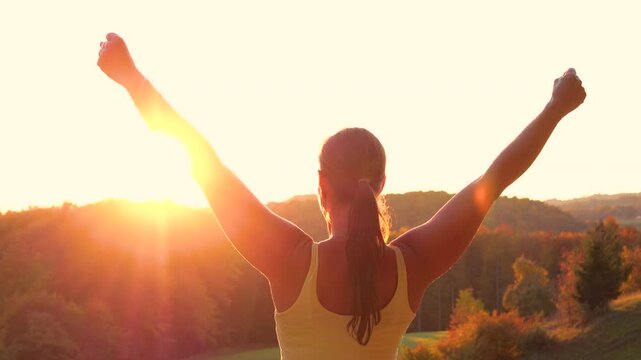 CLOSE UP, LENS FLARE: Woman on a scenic hill with both arms lifted in victorious pose. Warm golden sunlight and autumn colors spill over forested valley as she celebrates her sporting achievement.