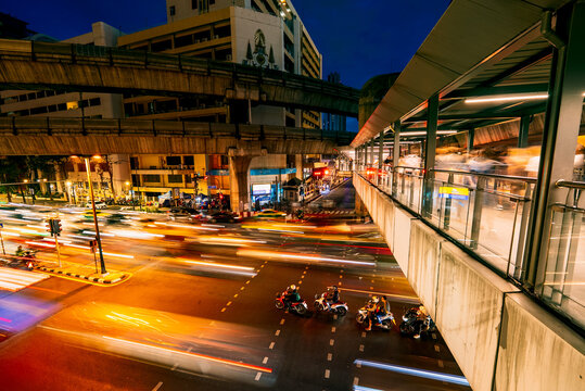 Bangkok, Bangkok, Thailand - 23 June 2024: View of a busy street intersection at night with vibrant light trails from vehicles, motorbikes, and a pedestrian walkway.