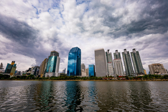 Bangkok, Thailand - 23 June 2024: View of the modern Khlong Toei skyline with glass skyscrapers and high-rise office buildings reflecting in the calm waters of Benjakitti Park under dramatic clouds.