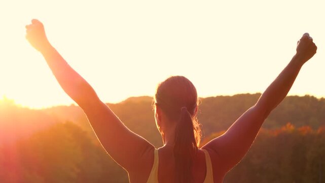 CLOSE UP, LENS FLARE: Young woman on a hilltop with her arms raised victoriously against glowing sunlight. Powerful, triumphant moment after morning run, framed by autumn hills and warm golden light.