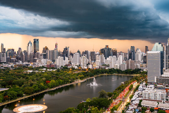 Bangkok, Thailand - 20 June 2024: View of Lumphini Park with its central lake and fountain, set against the modern skyline of Bangkok under a dramatic stormy sky.