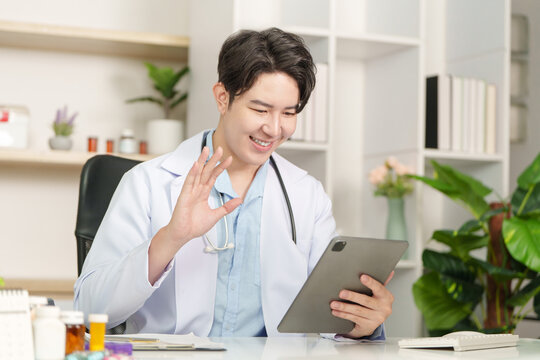 Asian male doctor waving on video call using digital tablet for telehealth consultation