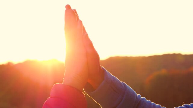 CLOSE UP, LENS FLARE, SILHOUETTE: Two adult hands meeting in a high five with the golden sun glowing behind them. Celebration, connection and shared success after an autumn hike in hilly countryside.