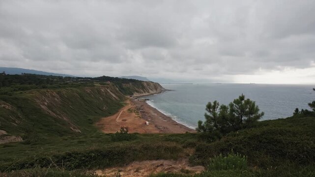 Vast coastal landscape featuring a sandy beach, eroding cliffs, and the Cantabrian Sea under an overcast sky, with distant ships