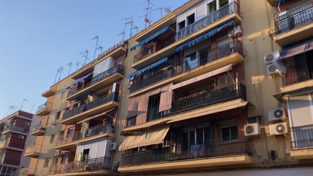 Row of urban residential buildings featuring balconies, colorful awnings, and numerous air conditioning units under a clear sky