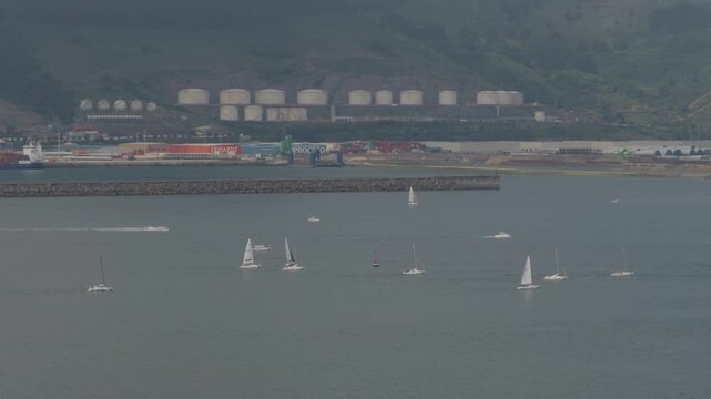 Sailboats cruising tranquil waters with an industrial port and green hills in the background, showing coastal lifestyle and travel