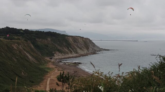 Paragliders enjoying views of a rugged coastline with cliffs, a beach, and the ocean under an overcast sky