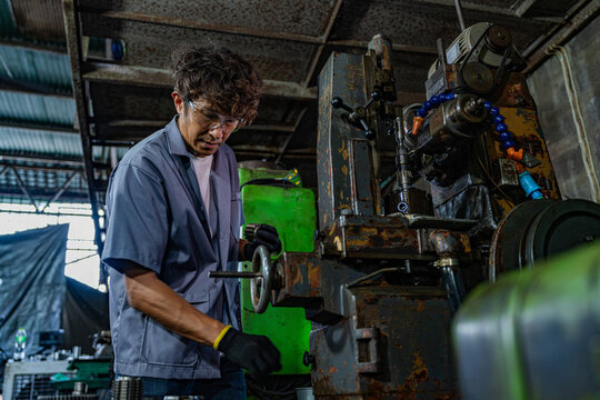 Middle aged Asian small business owner at his traditional machining workshop, dad's head bowed low, both gloved hands lowered to the handwheel/lead screw, expertise built over decades