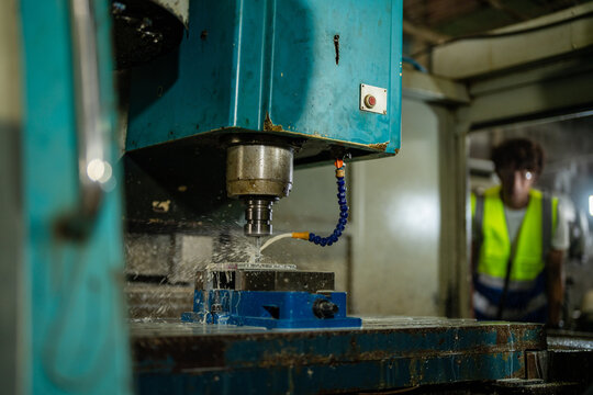 Asian father with curly hair inside his old family machine shop, dad stands upright across the machine, arms at his sides, watching the spindle, craftsmanship and dedication behind a small