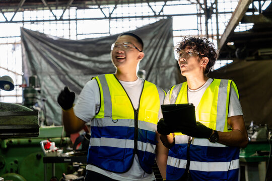 Senior craftsman father and his teenage son in a vintage family metal workshop, son's arm now lowered to shoulder height in an open-hand gesture, both smile, small family business heritage