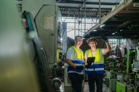 Middle aged Asian dad and teenage boy in a traditional metalwork factory, dad's arm still raised high pointing overhead, fingers spread slightly, family business succession and skill