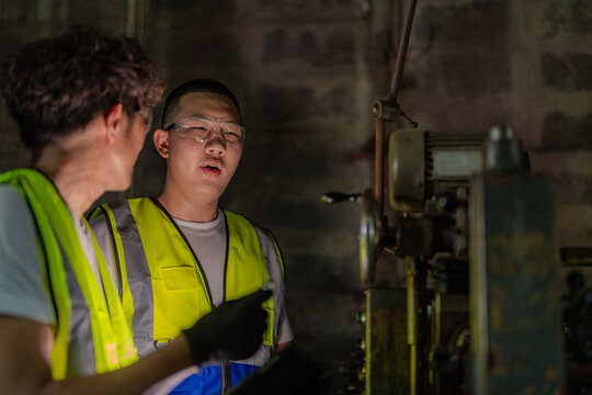 Thai father with curly hair and his teen son in a traditional metalwork factory, son talks and gestures with a gloved hand raised at his chest, dad in left, family business succession