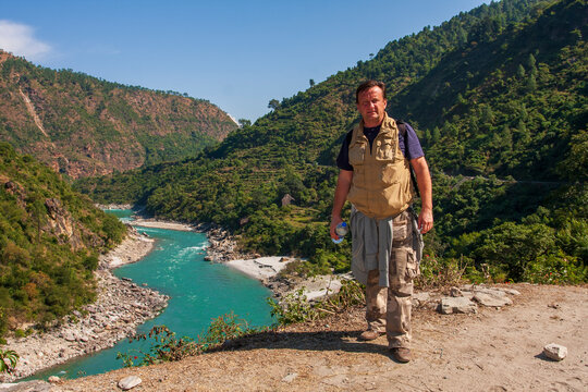 Man tourist in hiking clothes with backpack standing on viewpoint overlooking beautiful turquoise mountain river winding through green hills, India. Male traveler near mountain river valley