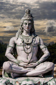 Statue of Hindu Lord Shiva meditating in lotus position, closeup. Lord Shiva statue in deep meditation post against cloudy dramatic sunset sky background in Rishikesh, India