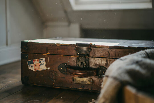 Antique wooden suitcase with a leather handle and vintage travel stickers placed on a dark wooden floor inside a rustic attic interior