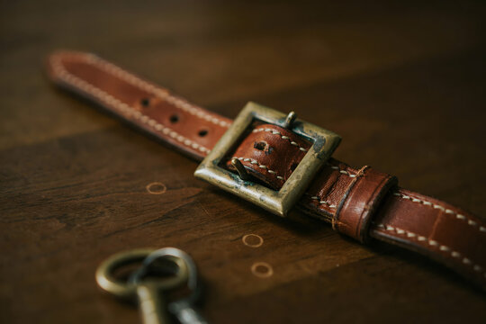 Brown leather watch strap with a brass buckle and vintage keys lying on a dark wooden surface under soft warm natural side lighting effects