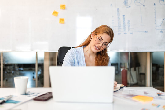 Business technology. Office laptop work, Woman looking at computer screen, Corporate and productivity concept. Smiling female professional sitting at desk with notebook and documents in modern office.