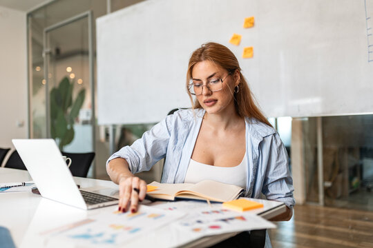 Business analysis. Office paperwork, Woman examining financial charts at desk, Corporate and data concept. Focused female professional reviewing documents and laptop in modern business interior.