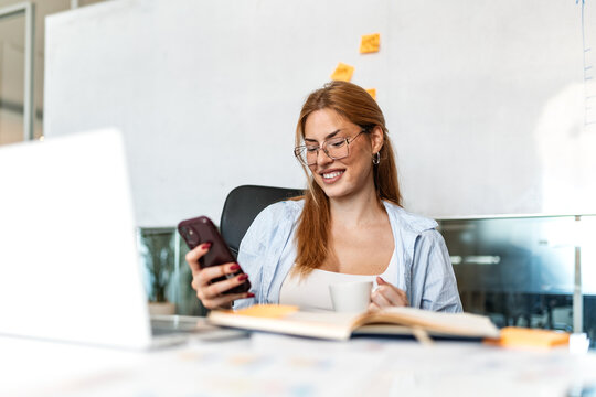 Business break. Office mobile message, Smiling woman holding smartphone and cup at desk, Corporate and tech concept. Happy female employee checking phone during work break in modern workplace.