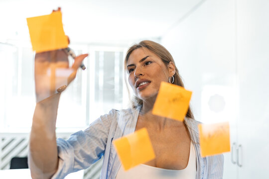 Project planning. Agile methodology, Woman writing on sticky notes glass board, Corporate management concept. Focused female professional organizing tasks with yellow post it notes in modern office.