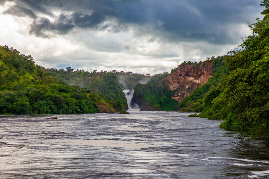 View of Murchison Falls on the Nile River surrounded by lush green vegetation and rocky cliffs under a dramatic cloudy sky in Uganda.