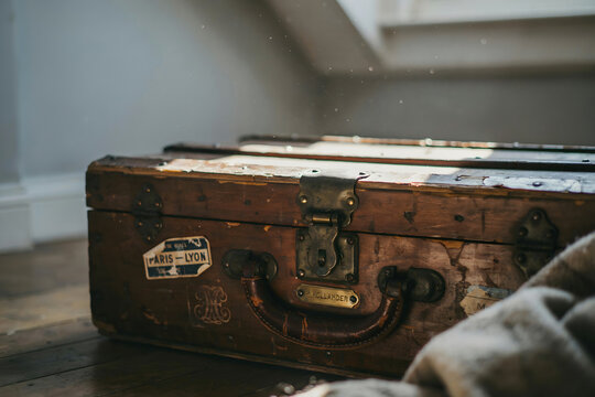 Old wooden travel trunk with metal hinges and worn paper labels sitting on a dark floor in a dimly lit room with soft window lighting