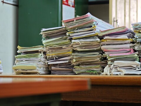 Massive stacks of old case files tied with strings sit on a wooden table inside a traditional government court room