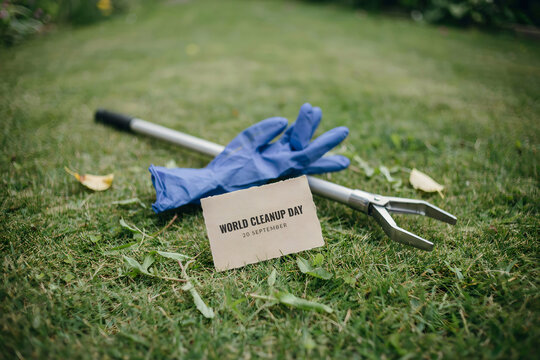 Metal trash picker tool and blue rubber gloves on green grass with a small card for cleanup day observed in the month of september outdoors