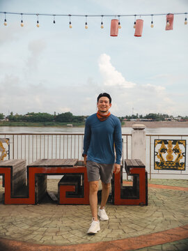 Happy young Asian man enjoying a walk along the river promenade with decorative lights.