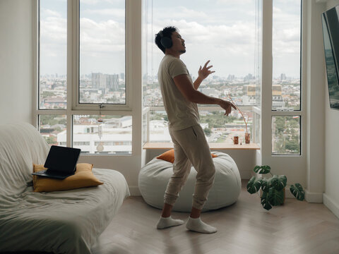 Energetic young man wearing headphones, dancing and singing freely in his bright city apartment.