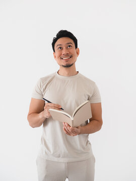 Smiling Asian man holding a notebook and pen, looking up thoughtfully, getting an idea.