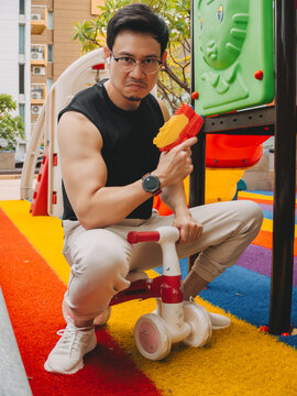 Man playfully squats on a balance bike, holding a toy water gun at the playground.