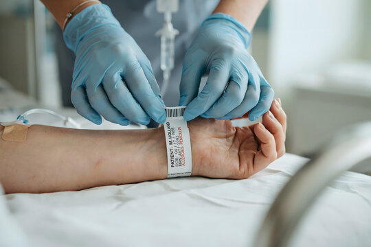 A medical wearing blue gloves secures a patient identification wristband on the arm of a person lying in a hospital bed with iv drip
