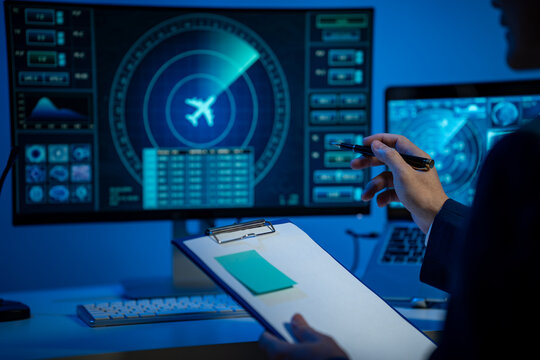 Person in a dark office pointing at a screen with an airplane dashboard and holding a clipboard, showing aviation monitoring and data analysis in blue light