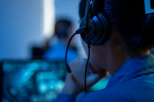 Close-up of a call center worker wearing a headset in front of blurred computer screens, with blue office lighting and a busy support team in the background