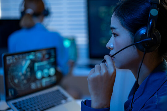 Woman wearing a headset and microphone working at a computer in a dim office with blue light, while another person sits blurred in the background