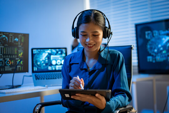Young asian woman wearing a headset uses a tablet in a modern office with multiple screens, suggesting online support, planning, and digital work.