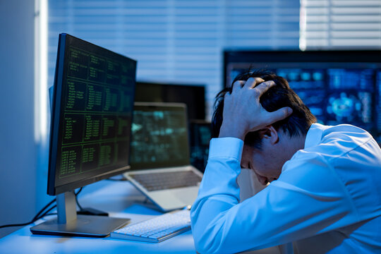 Stressed office worker sitting at a desk with computer screens, holding his head in frustration after long hours of work