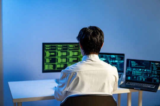 Rear view of a person working at a desk with multiple computer monitors showing code and data in a dark office