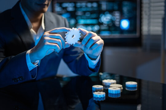 Businessman holding a white gear with blue lights in a modern office, with other gears on a table and a blurred technology screen in the background.