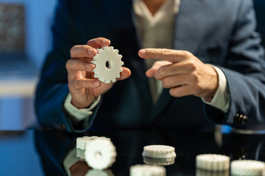 Man in a business suit holding a white gear while pointing at it during a technical discussion, with several gears on a glossy table