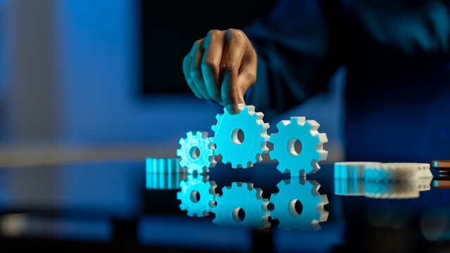 Hand placing a gear into a row of connected cogs under blue light, showing business teamwork, automation, and engineering concept.