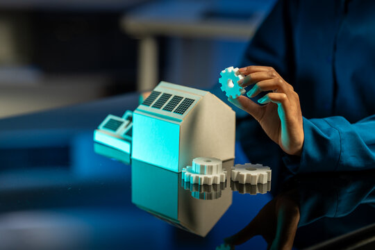 Hand placing a gear beside a miniature solar powered house on a reflective table, symbolizing clean energy, innovation and sustainable living.