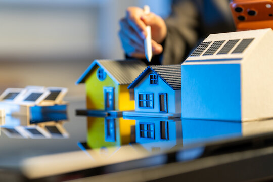 Miniature houses with solar panels on a reflective table while a hand points with a pen, showing home planning, real estate, and clean energy concept.
