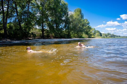 Two male friends swimming and splashing in a river with forested shoreline under a blue sky on a sunny summer day