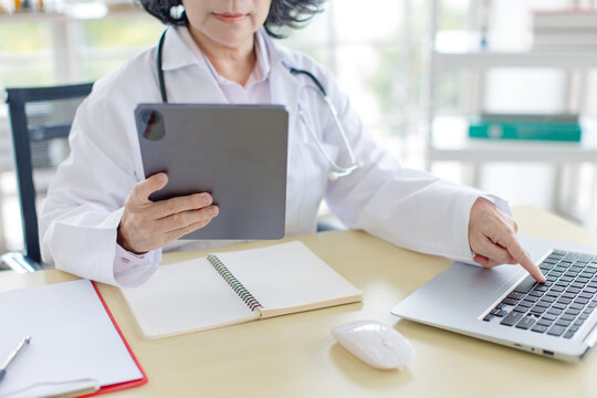 Asian smiling older woman doctor wearing medical coat stethoscope working reading data tablet laptop in healthcare office professional physician services hospital clinic wellness trust patient care.