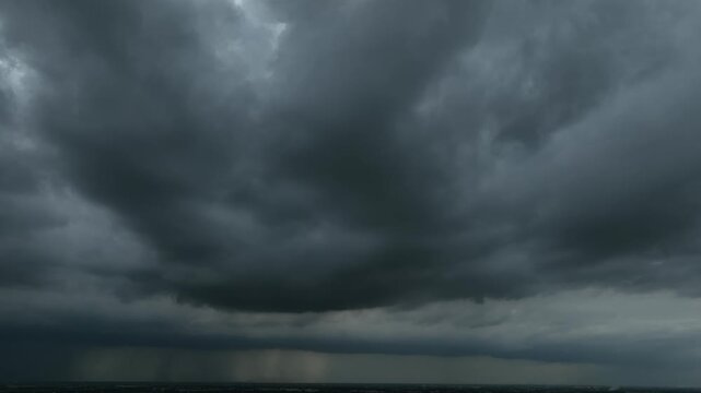 A massive storm clouds releasing heavy rain showers toward the landscape below, capturing dramatic dark formations, rainfall curtains, and the intense atmosphere of an approaching thunderstorm. 4k.

