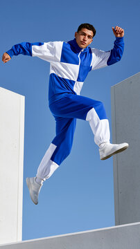 Athletic man in blue and white tracksuit captured mid-jump between concrete structures, dynamic parkour pose against clear blue sky, bold sportswear editorial fashion styling.