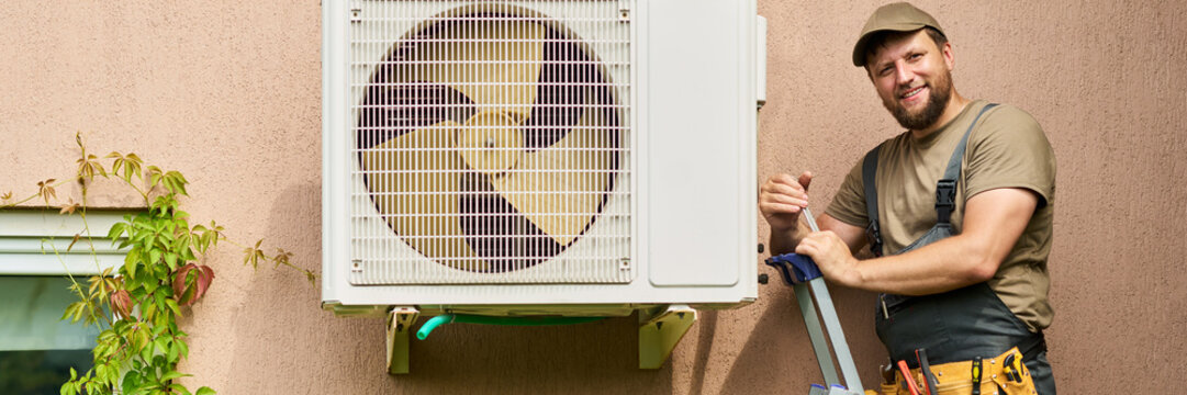 Portrait of Caucasian young adult male technician smiling while standing on ladder next to outdoor air conditioner unit, representing HVAC maintenance services, medium shot